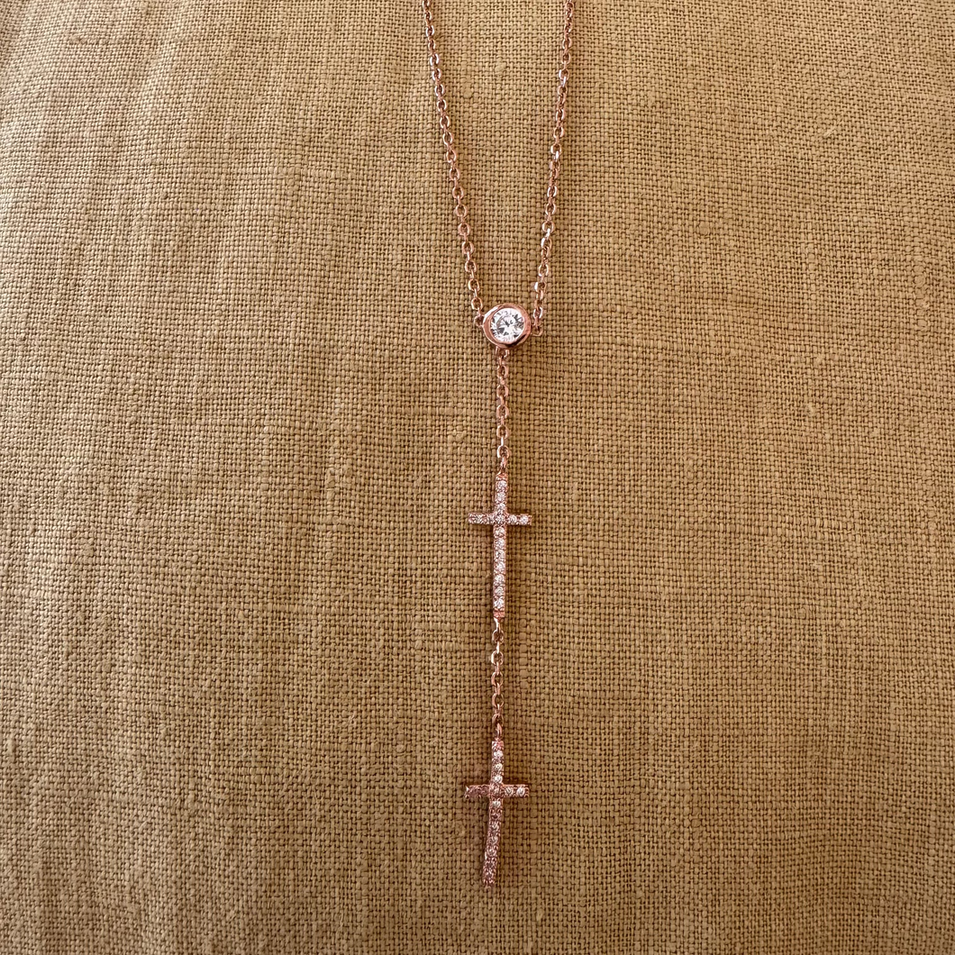 A rose gold-plated lariat necklace with cubic zirconia and two hanging crosses, displayed against a beige fabric background.