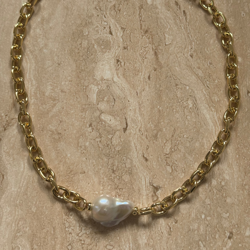 A gold-colored chunky link chain bracelet featuring a large baroque fresh water pearl in the center, displayed on a marble surface.