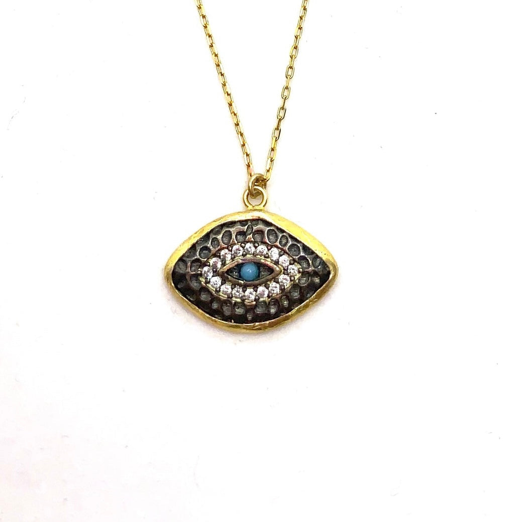 A turquoise mati-eye necklace with oxidised details, yellow and rose gold plating, and sterling silver, displayed against a beach backdrop.