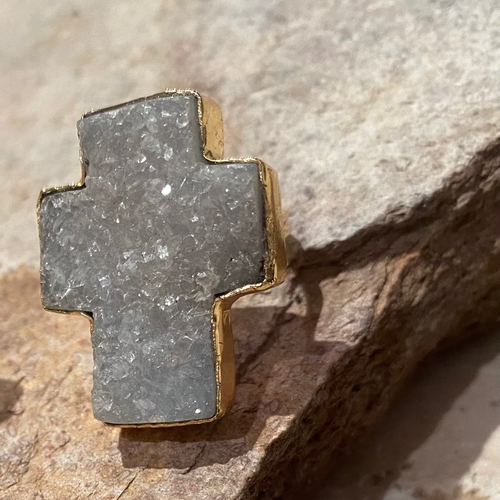 A gold-plated sterling silver ring with a large cross design, featuring a light blue pink cracked quartz center, placed on a textured surface.