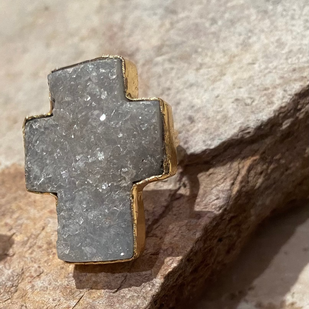 A gold-plated sterling silver ring with a large cross design, featuring a light blue pink cracked quartz center, placed on a textured surface.