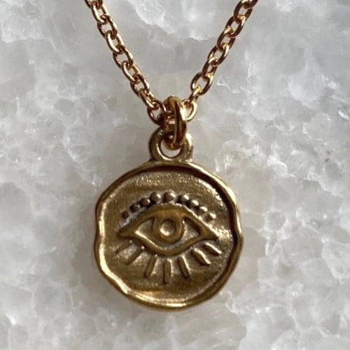 A gold vermeil coin necklace with an evil eye design on a chain, displayed on a textured background.