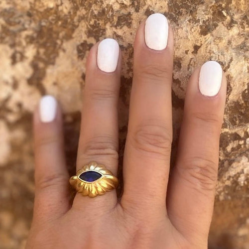 A close-up image of a woman's hand with a white manicure, featuring a gold vermeil and blue cubic zirconia ring on the finger.