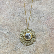 A silver necklace featuring a crocheted round design with a coin depiction of the goddess Athena in the center, attached to a gold vermeil chain, displayed against a stone background.